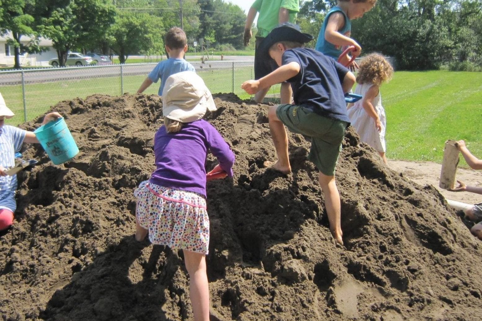 Nine children playing outside on and around a large pile of sand.